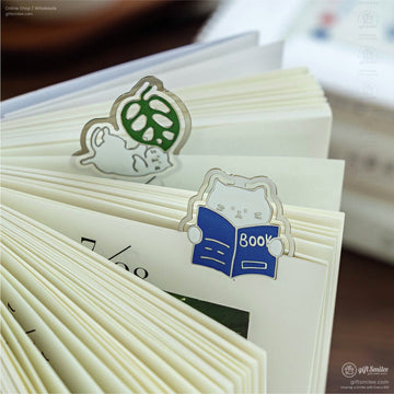 Close-up of cute cat-shaped enamel bookmarks clipped onto book pages.