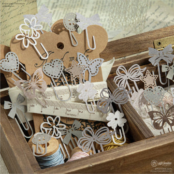 A wooden box filled with vintage-style decorative paper clips, including butterfly, bow, and floral designs