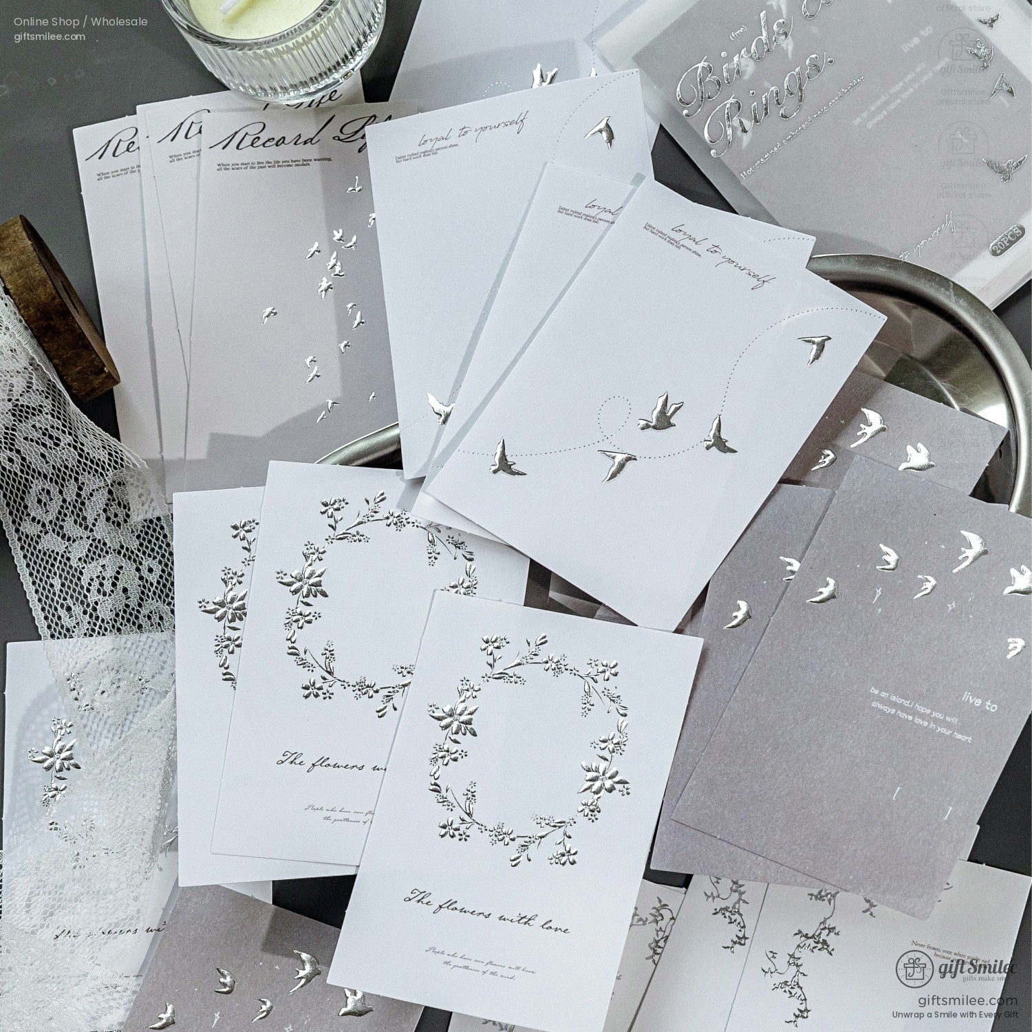 A detailed set of silver foil stationery cards with bird, floral, and wreath illustrations on a dark surface