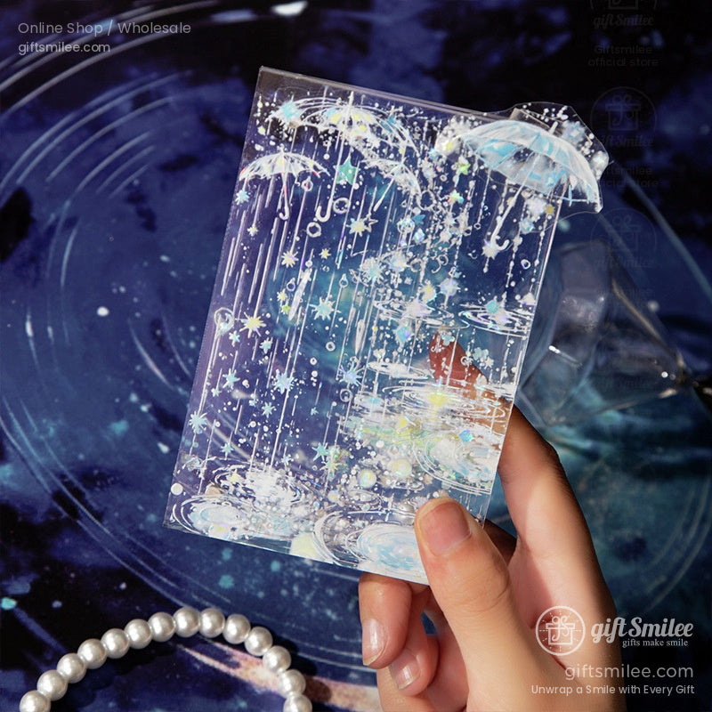 A hand holding translucent washi tape showcasing sparkling rain and umbrellas, set against a cosmic background